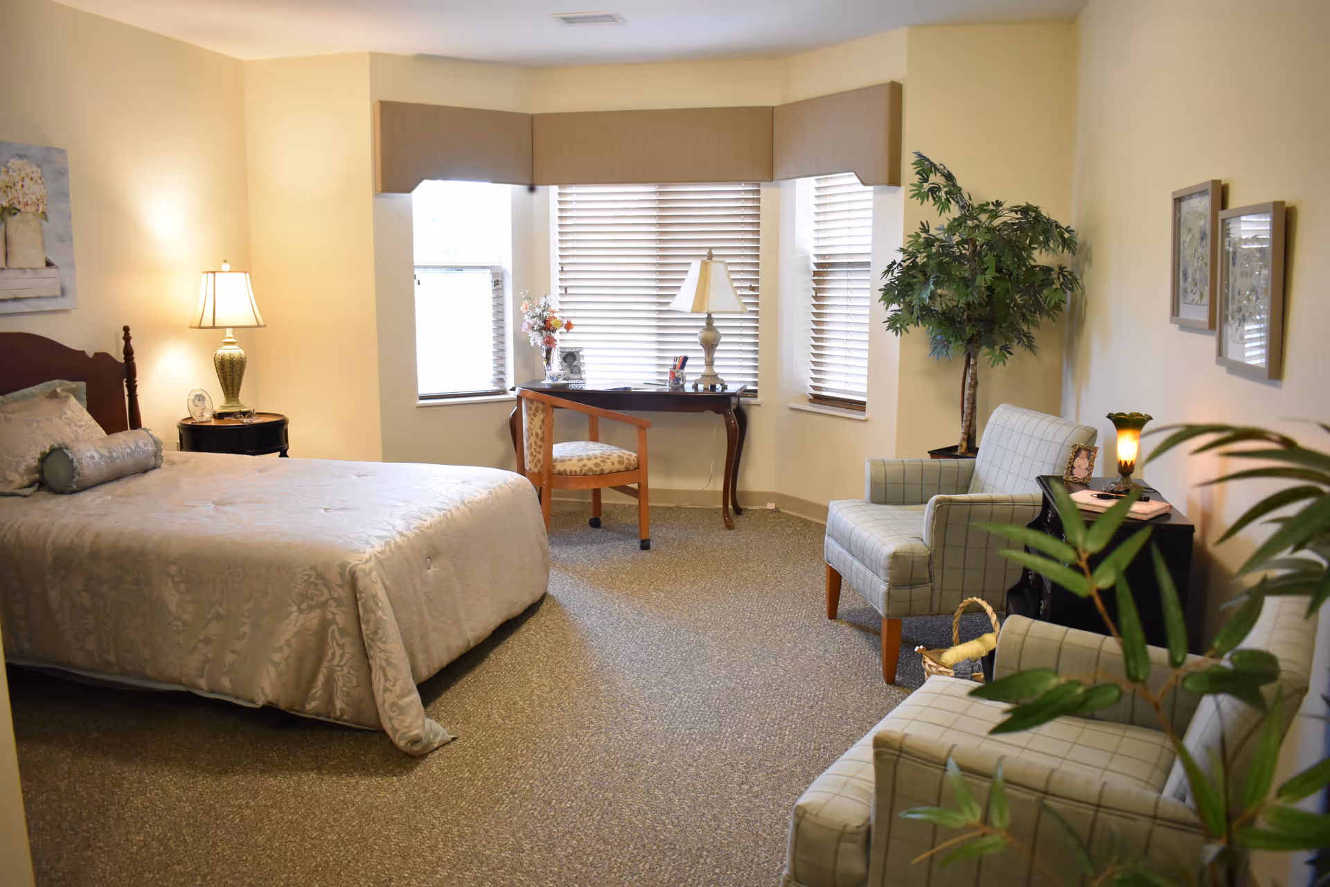 A cozy bedroom with a large bed covered in a beige patterned bedspread, a wooden nightstand with a lamp, a small desk with a chair in front of three windows with blinds, two upholstered armchairs, a side table with a lamp, and a potted plant in the corner.