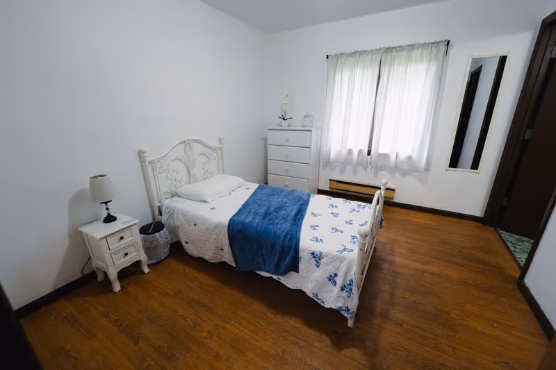 A simple bedroom with a single white metal bed frame, a white nightstand with a lamp, a white chest of drawers, a window with white curtains, and wooden flooring.