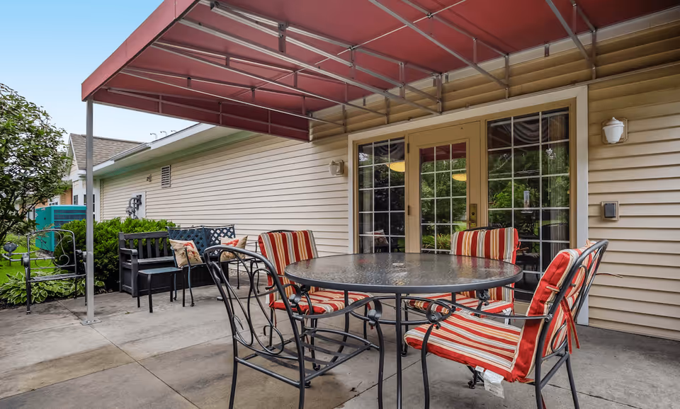 Outdoor patio area with a round glass table and four metal chairs with red and white striped cushions. The patio is covered by a red awning attached to a beige siding building with glass double doors. There are additional benches and greenery along the side of the building.