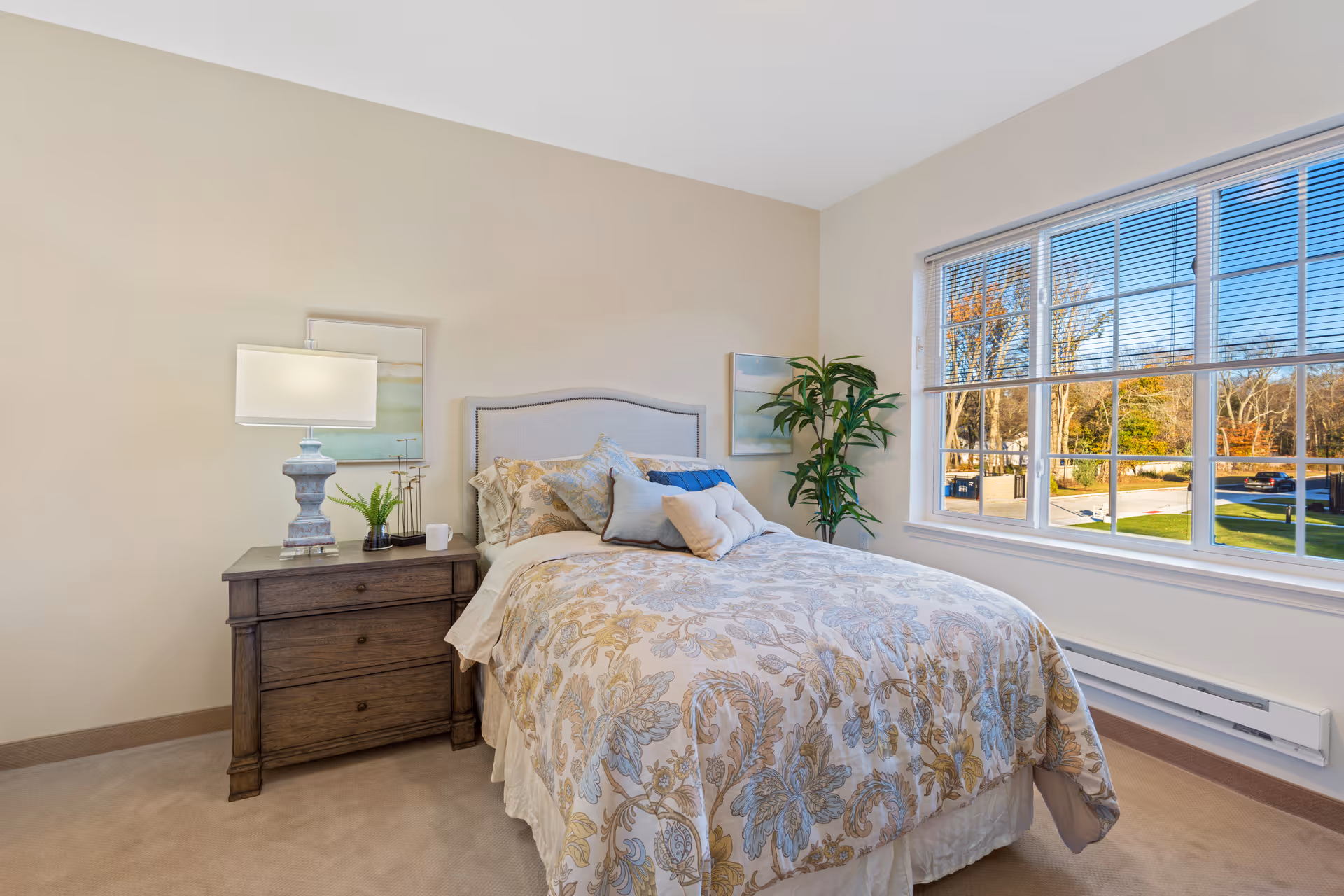 A cozy bedroom with a neatly made bed featuring a floral patterned comforter and multiple pillows. Next to the bed is a wooden nightstand with a lamp, a small plant, and a cup. The room has beige walls and a large window with blinds, showing a view of trees and a parking area outside.