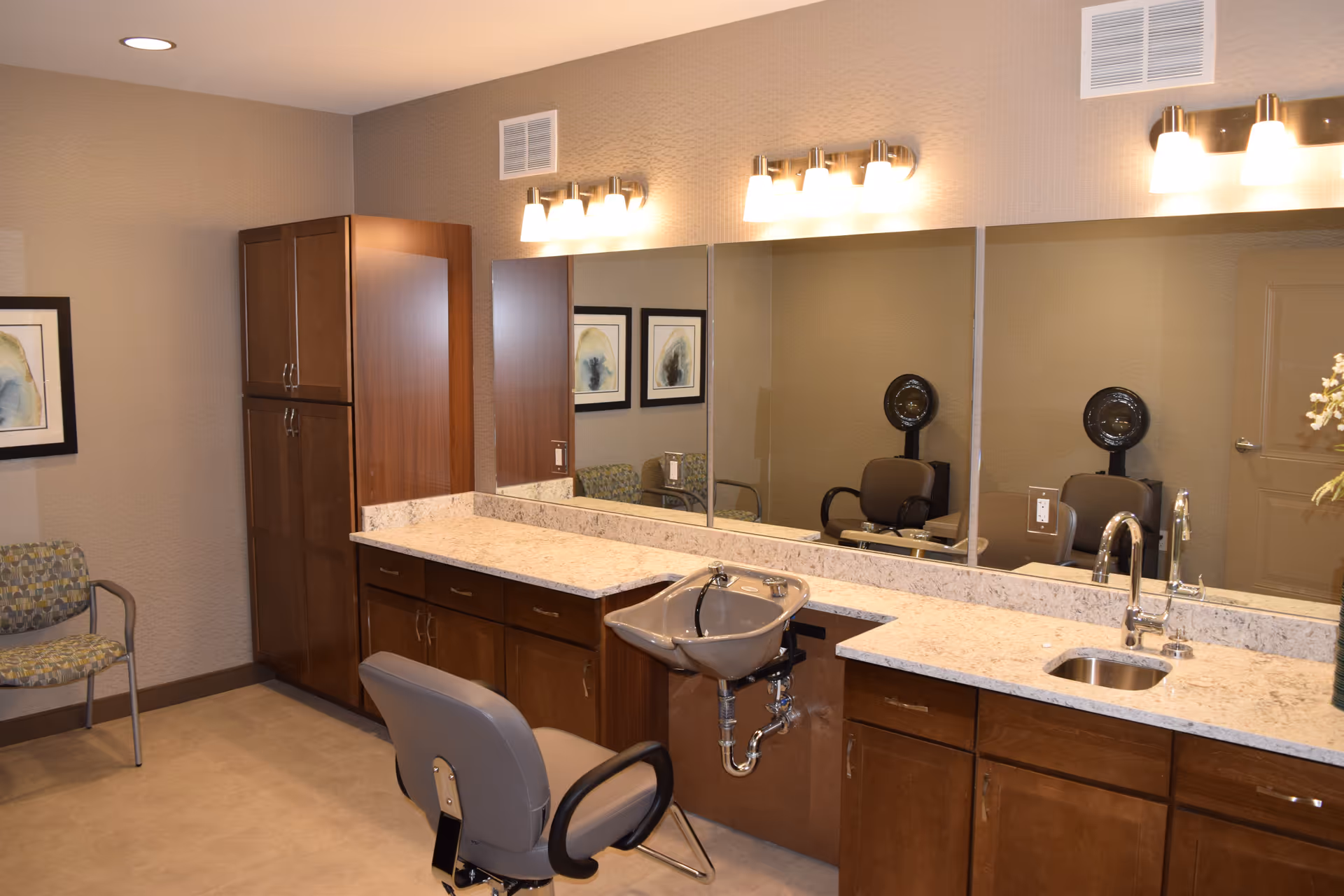 Interior view of a salon area in an assisted living facility featuring a salon chair in front of a countertop with a built-in hair washing sink, large mirrors, wooden cabinets, and wall-mounted light fixtures. There is also a patterned chair and framed artwork on the wall.