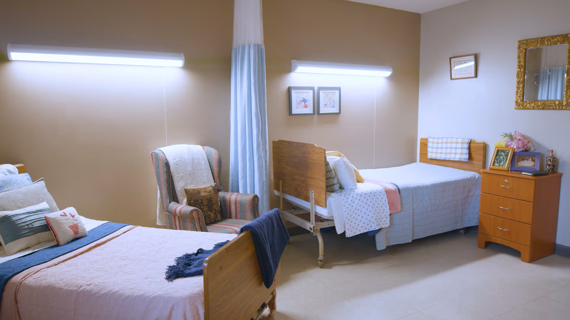 Two beds in a senior care room with an armchair, bedside dresser and overhead lighting.