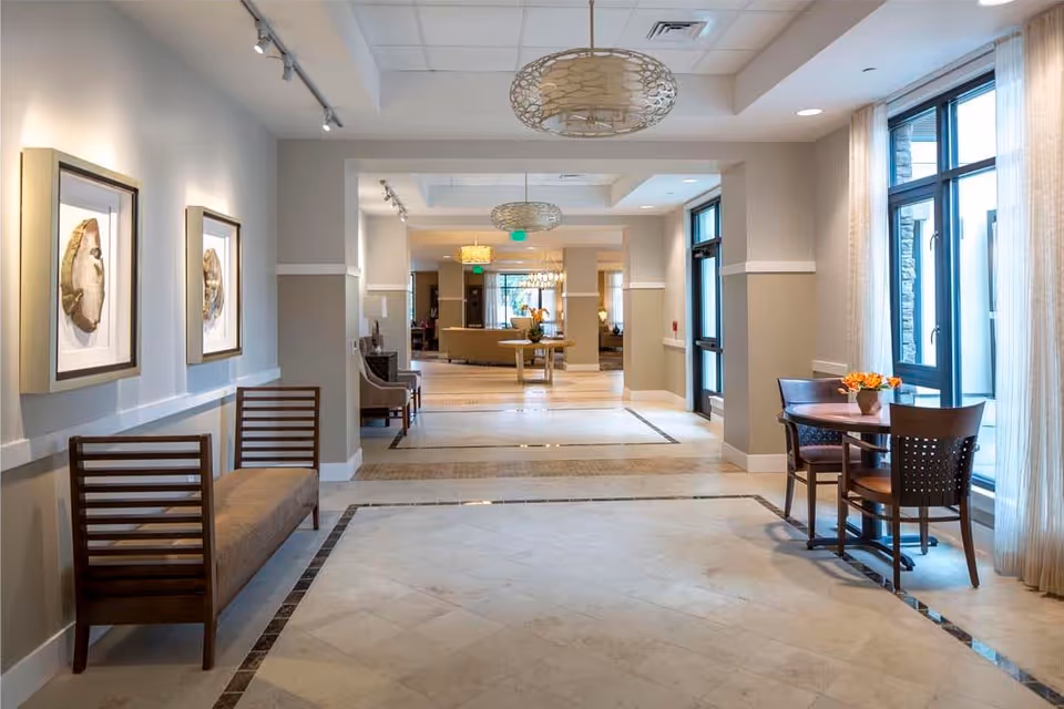 A bright and spacious hallway in a senior living facility with beige walls and tiled floors. On the left side, there are two framed artworks above a wooden bench with a cushioned seat. On the right side, there is a small round table with four chairs and a vase with orange flowers near large windows with sheer curtains. The hallway leads to a common area with more seating and decorative lighting fixtures hanging from the ceiling.