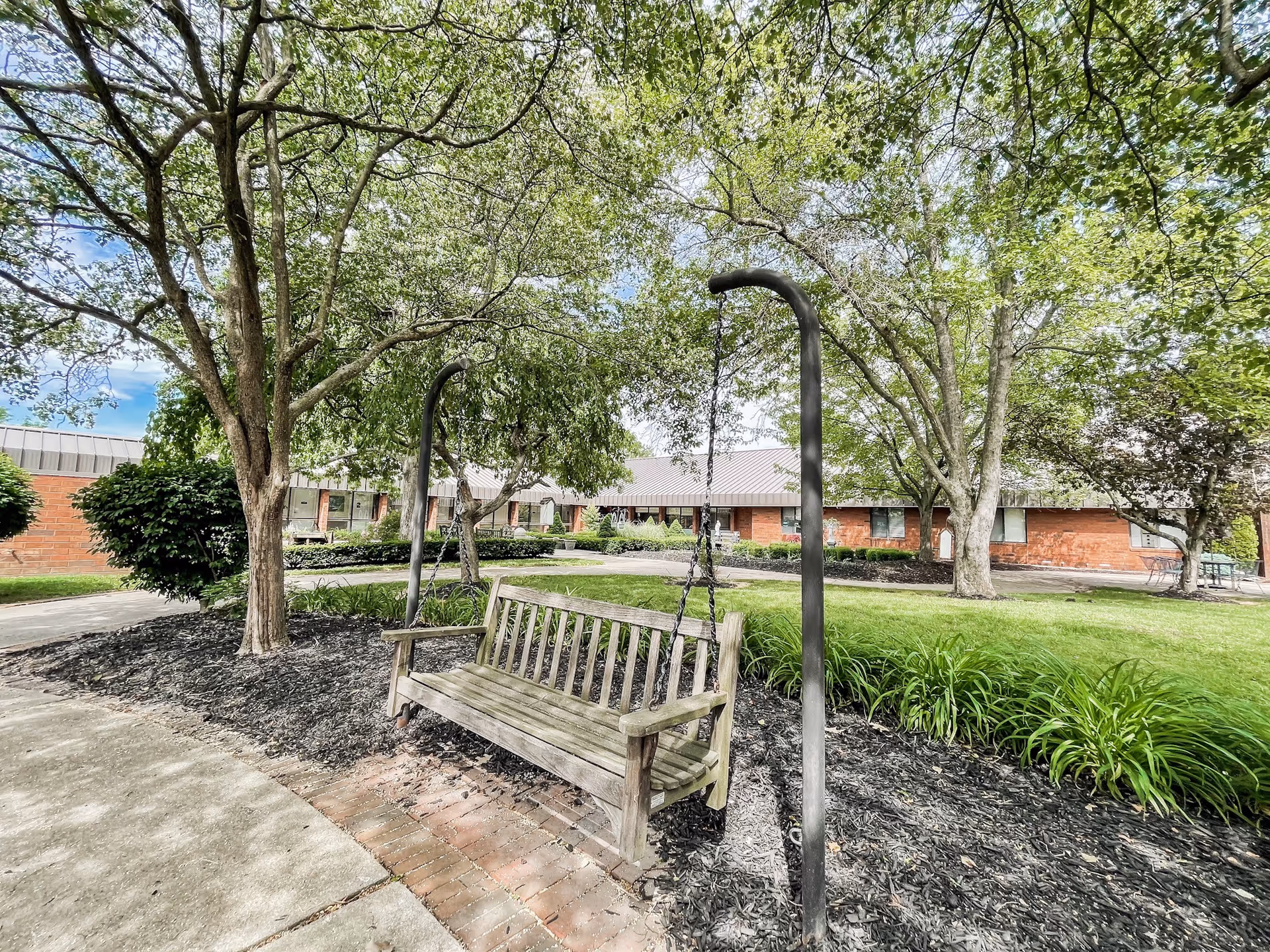 A peaceful outdoor garden area at Inniswood Health & Rehabilitation featuring a wooden swing bench suspended by chains between two metal posts, surrounded by trees, green grass, and landscaped plants with a brick building in the background under a partly cloudy sky.