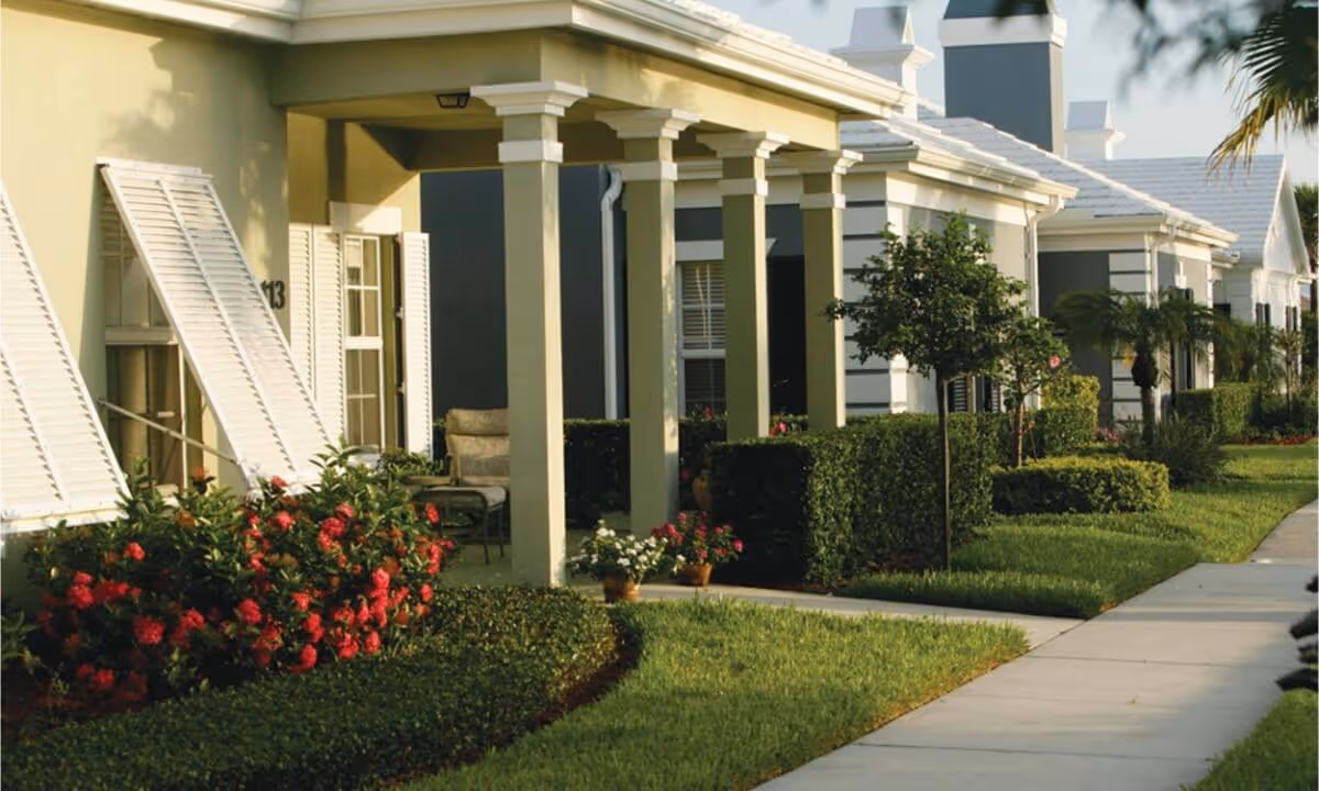 Row of single-story senior living units with covered porches, columns, manicured landscaping and a sidewalk.
