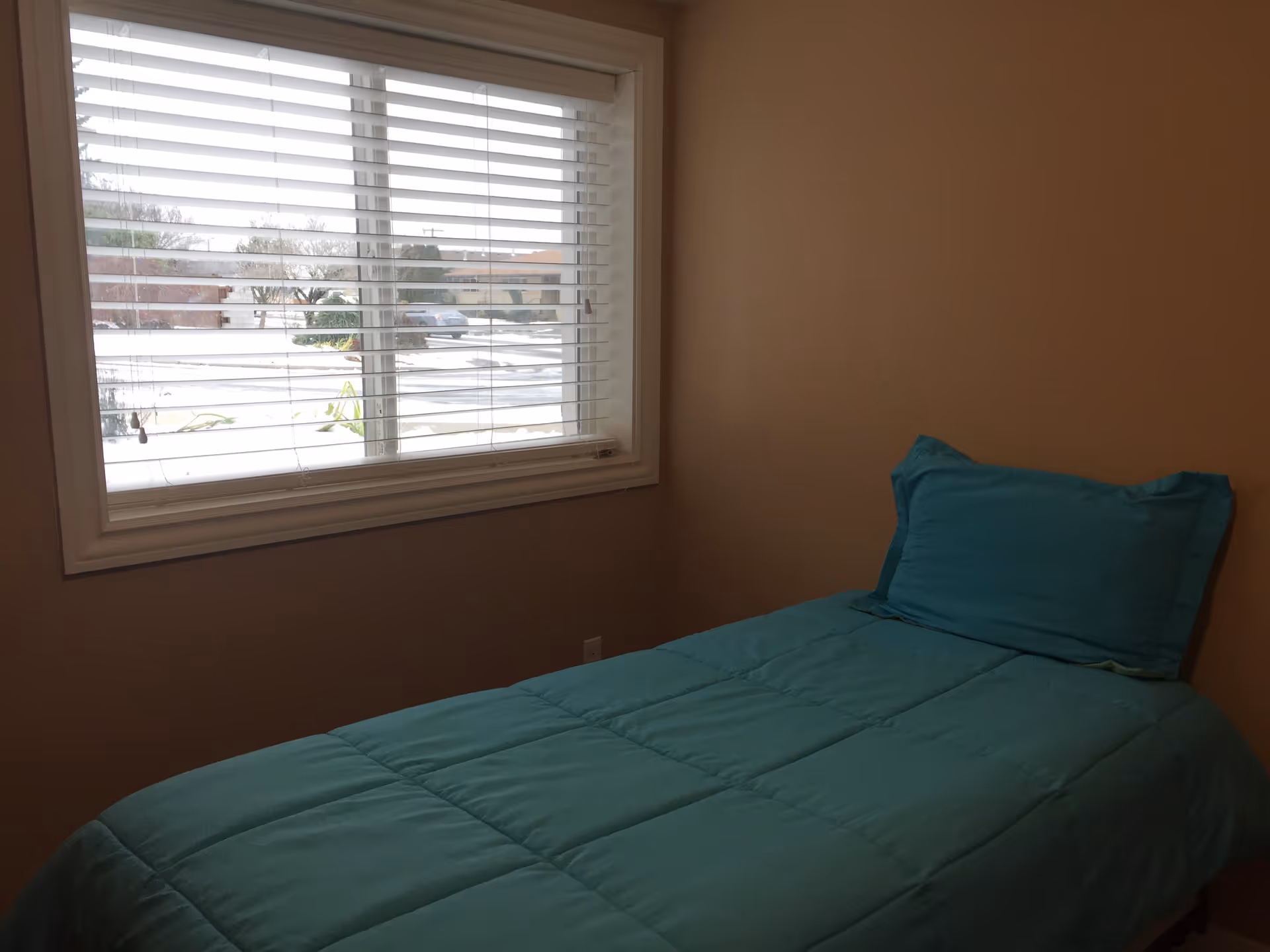 A small bedroom with beige walls, a window with white blinds, and a single bed with a teal comforter and matching pillow.