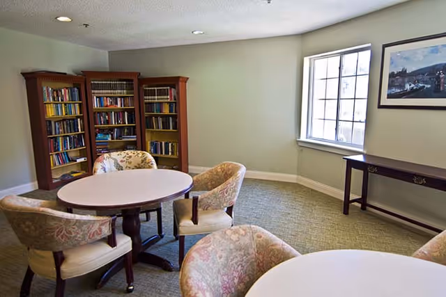 A cozy room with round tables surrounded by floral upholstered chairs, a wooden bookshelf filled with books, a window letting in natural light, and a framed picture on the wall.
