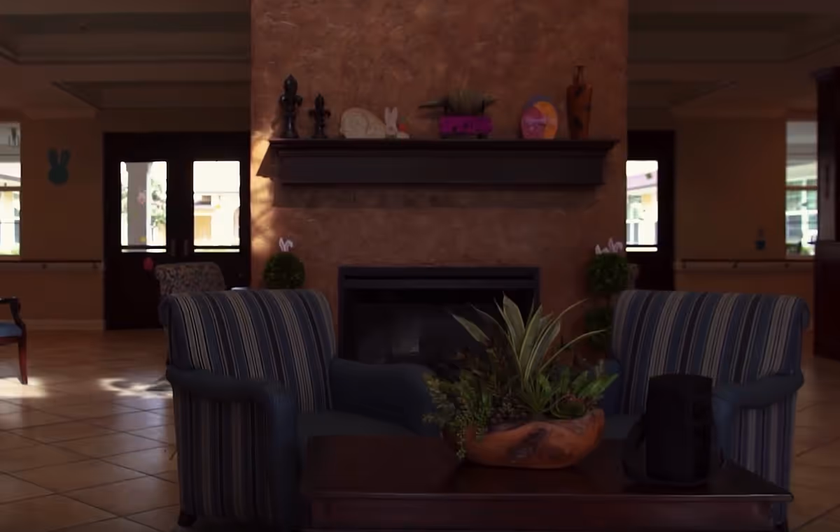 A cozy seating area with two striped armchairs facing a wooden coffee table that holds a decorative plant and a black object. Behind the seating area is a fireplace with a mantel decorated with various ornaments including vases and figurines. The room has tiled flooring and large windows letting in natural light.