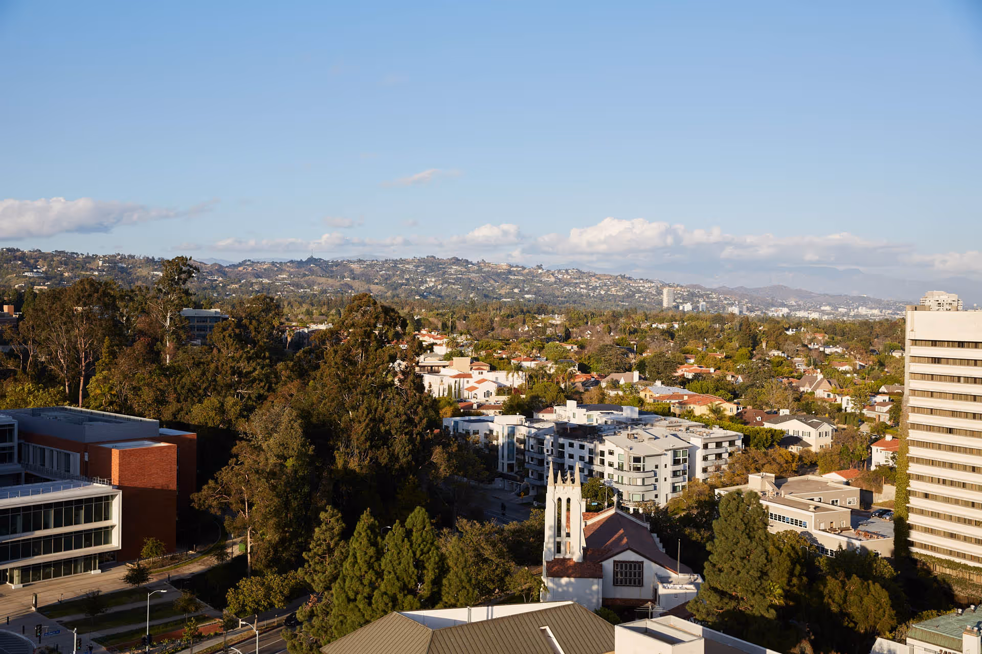A wide aerial view of a suburban area with a mix of residential houses, apartment buildings, and trees under a clear blue sky with some clouds. Hills are visible in the background.