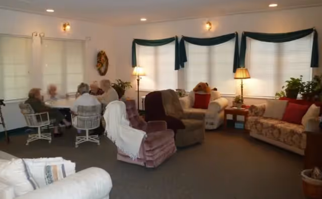A cozy living room in an assisted living facility with several armchairs and sofas arranged around the room. Four elderly people are seated at a round table near the windows, engaged in conversation. The room is softly lit with floor lamps and decorated with plants and cushions.