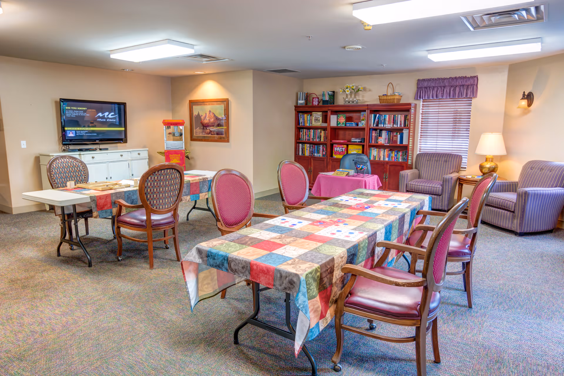 A cozy common room in a senior living facility with two tables covered in colorful patchwork tablecloths surrounded by wooden chairs with cushioned seats. In the background, there is a bookshelf filled with books, two striped armchairs, a side table with a lamp, a window with purple valance, and a wall-mounted TV above a white cabinet. The room has carpeted floors and soft lighting.