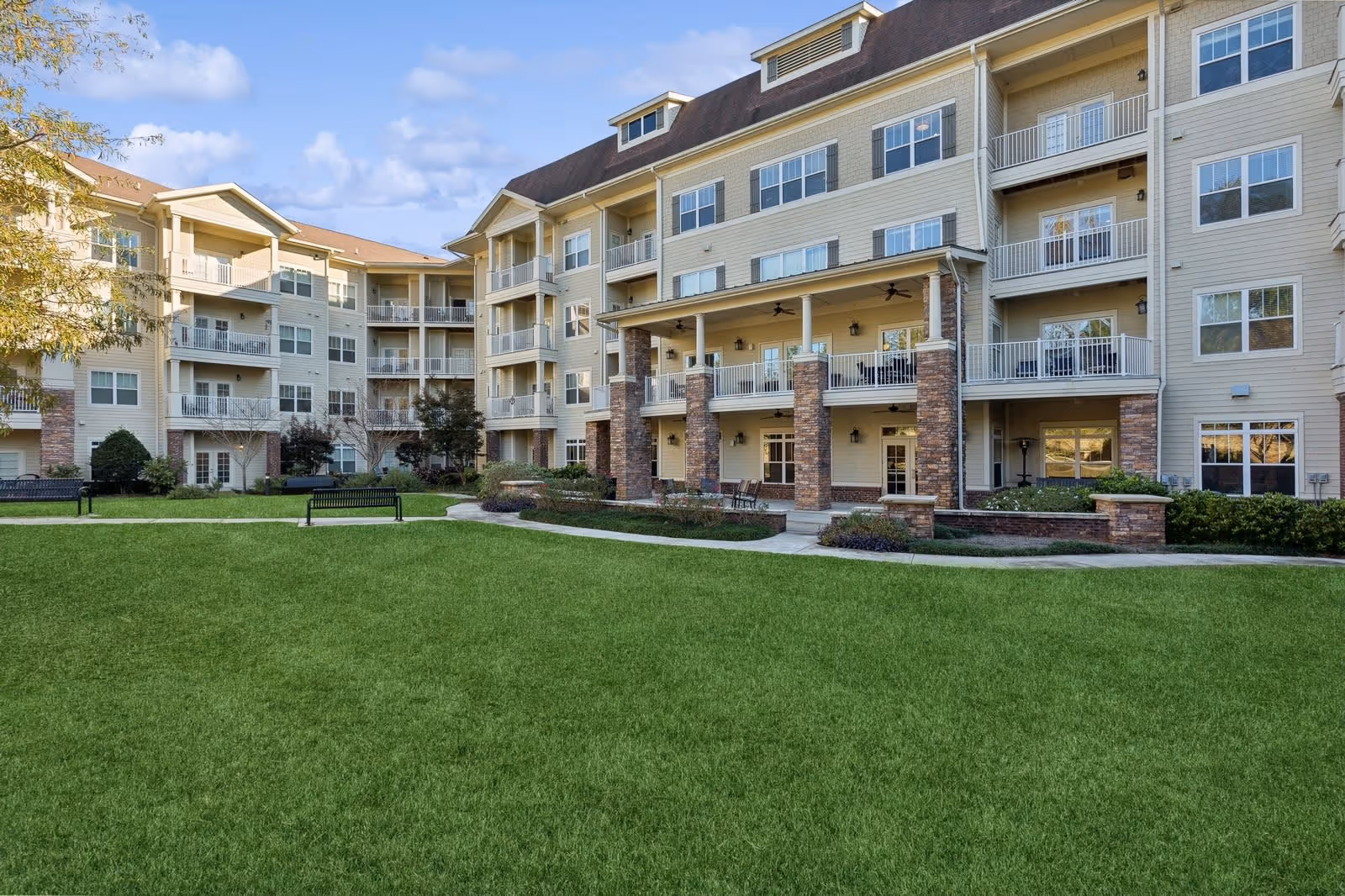 Exterior view of a multi-story senior living facility building with balconies and a well-maintained green lawn in the foreground under a partly cloudy sky.
