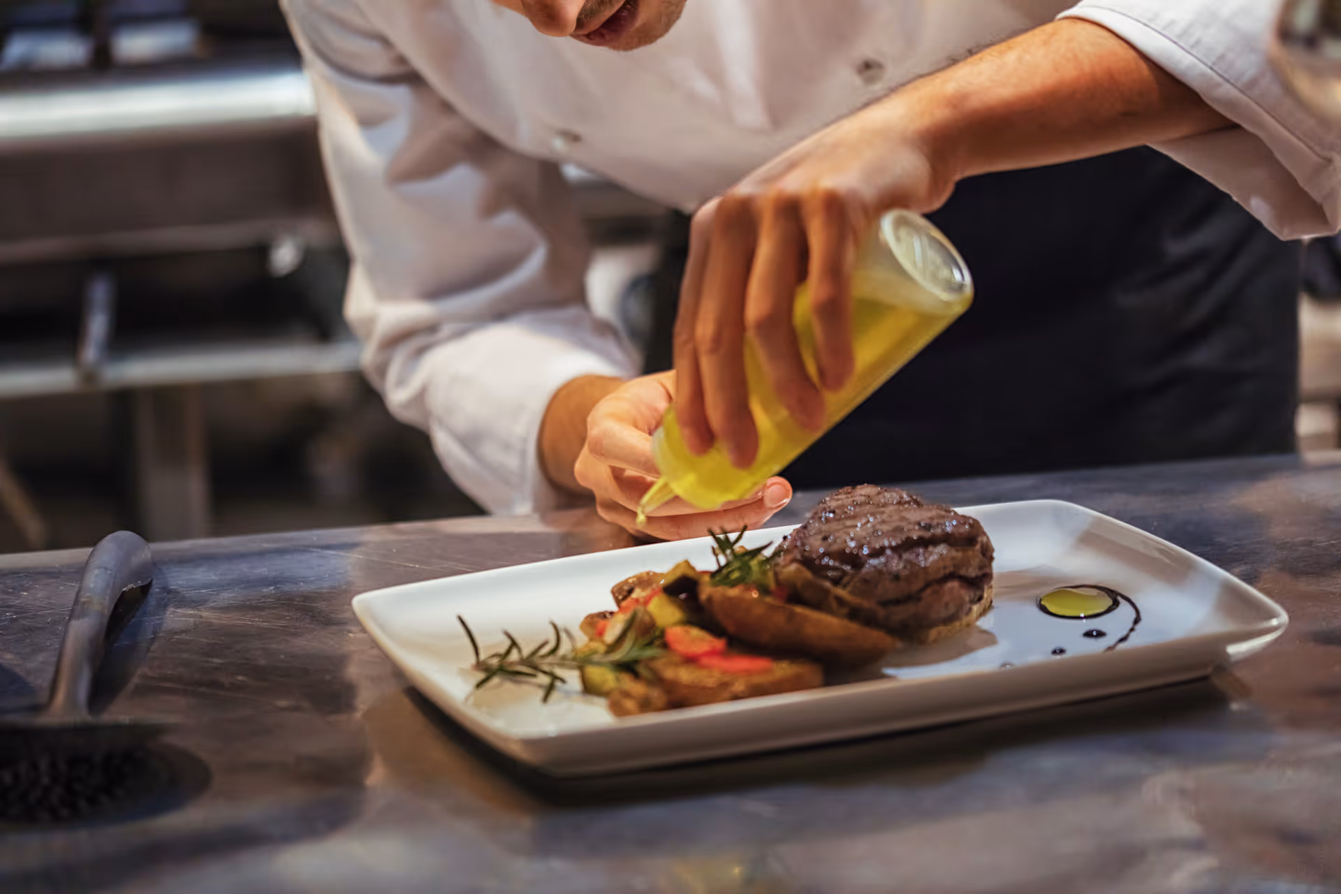 A chef in a white uniform is carefully drizzling a yellow sauce onto a plated dish that includes a grilled steak and roasted vegetables, garnished with herbs, on a white rectangular plate in a professional kitchen.
