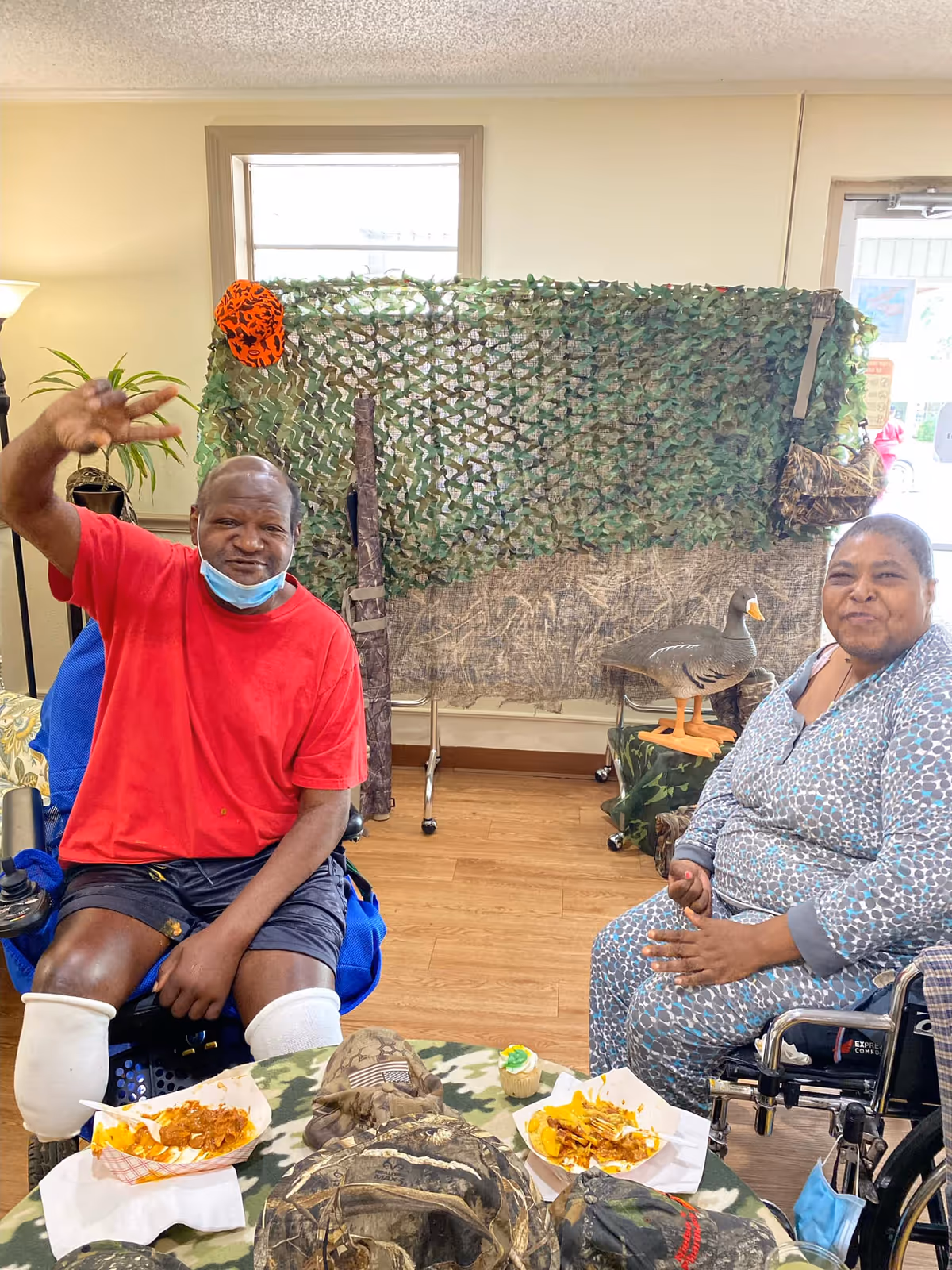 Two people sitting in wheelchairs inside a room with wooden flooring. One person is wearing a red shirt and black shorts with white leg braces, waving at the camera. The other person is wearing a patterned outfit and smiling. In front of them is a table with camouflage hats and plates of food. Behind them is a camouflage netting decoration with a fake duck on a stand and a window letting in natural light.