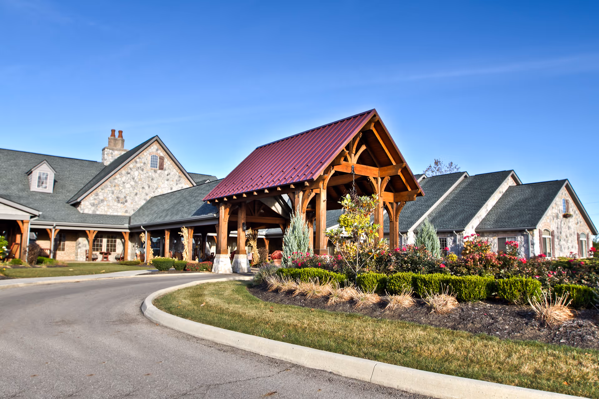 Front exterior of a stone-faced senior living facility with a wooden covered entrance, circular driveway, and landscaped flower beds under a clear blue sky.