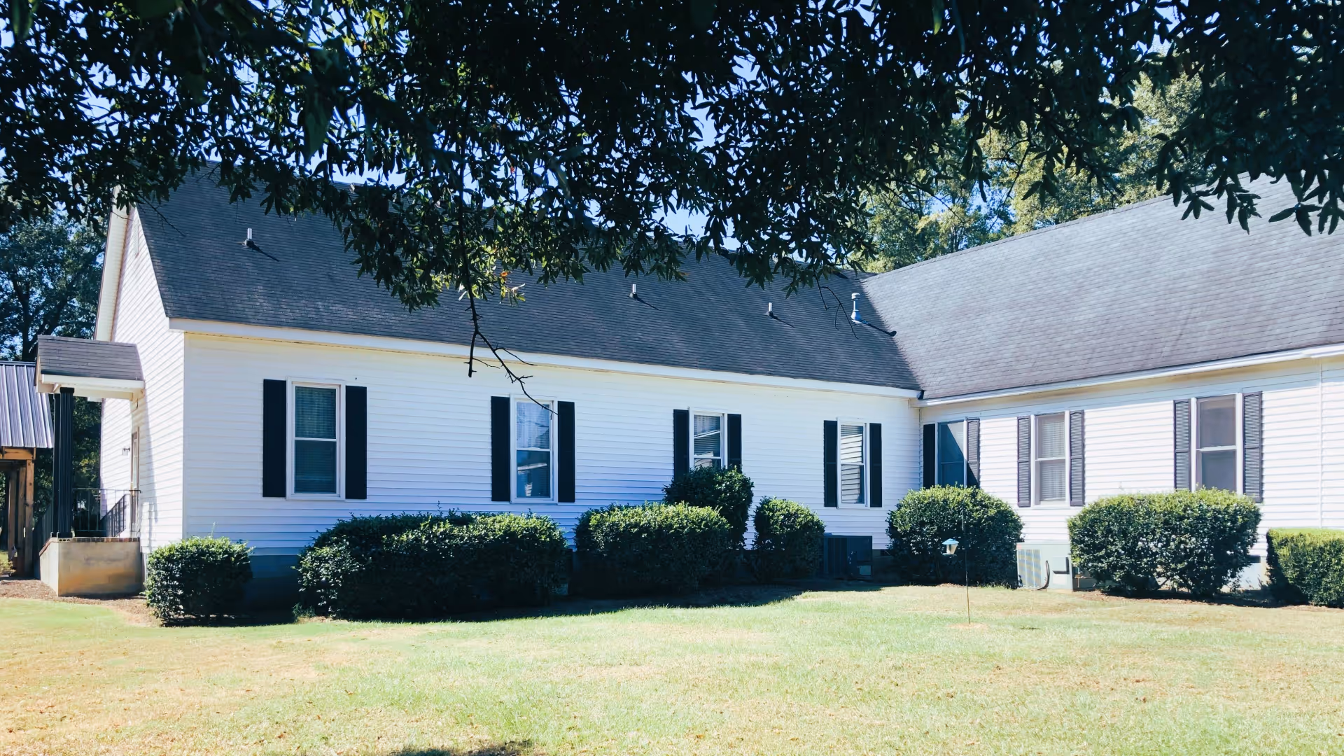 Single-story white siding building with black shutters, neatly trimmed bushes and a grassy lawn under overhanging tree branches.