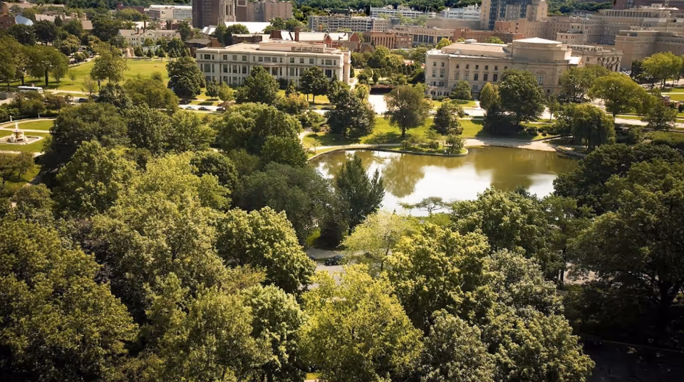 Aerial view of a park with dense green trees surrounding a pond, with several large buildings in the background under a clear sky.