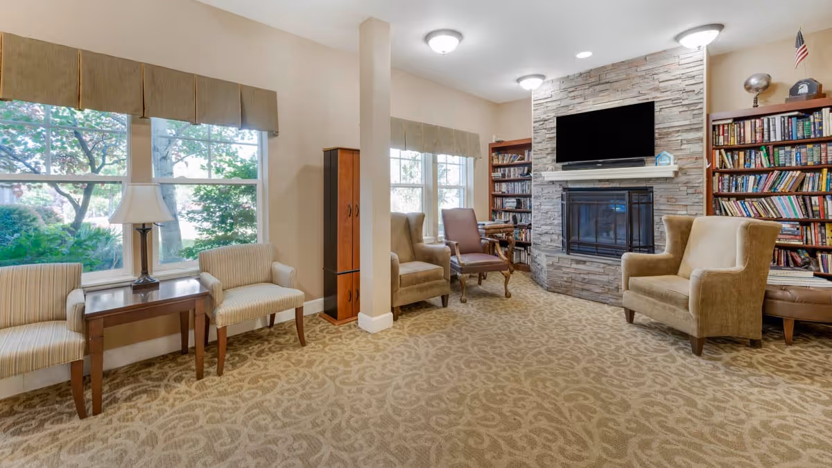 A cozy living room area with beige patterned carpet, several upholstered chairs, a wooden side table with a lamp, large windows with valances showing greenery outside, a stone fireplace with a mounted flat-screen TV above it, and bookshelves filled with books on either side of the fireplace.
