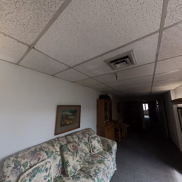 A dimly lit hallway in a retirement community with a floral patterned couch against a white wall, a framed picture above the couch, a wooden cabinet with glass doors, and a couple of wooden chairs further down the hallway. The ceiling has white acoustic tiles with a vent and a light fixture.