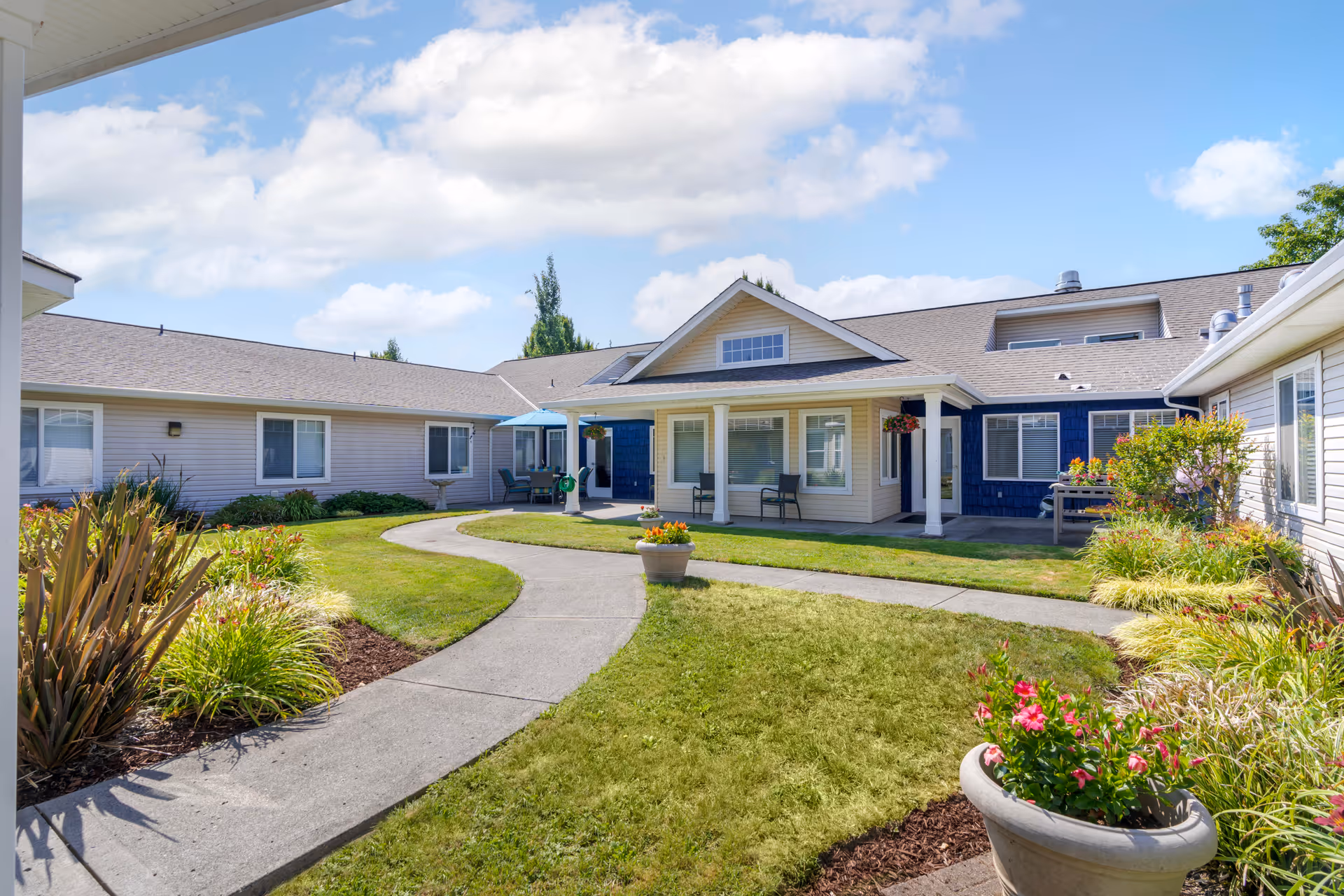 Outdoor courtyard area of a senior living facility with a curved concrete walkway, green grass, flower beds, and potted plants. The building surrounding the courtyard has beige and blue siding with multiple windows and a covered porch with chairs. The sky is partly cloudy and blue.