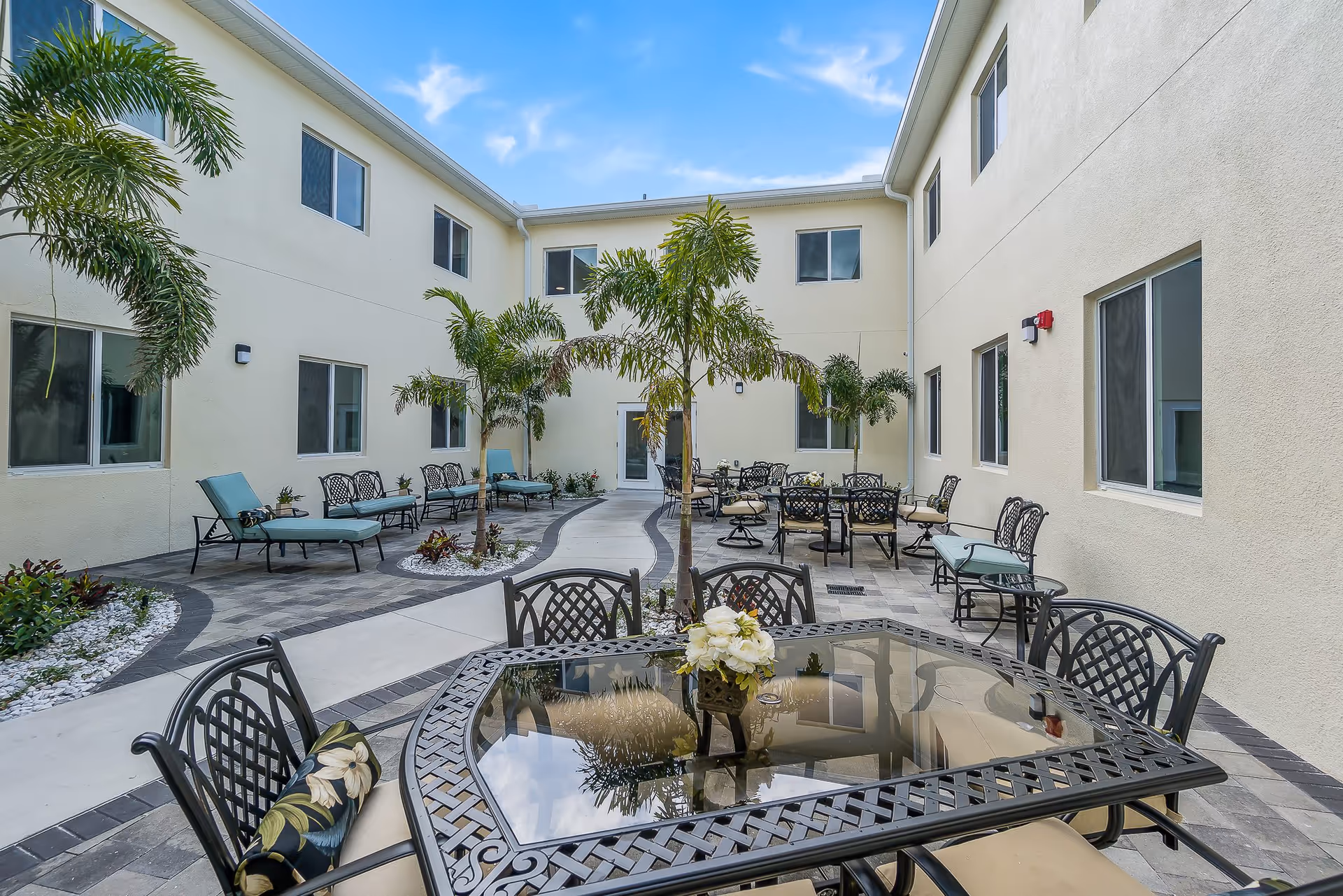 Sunlit courtyard with patio tables, chairs, lounge seating and palm trees surrounded by a two-story beige building.