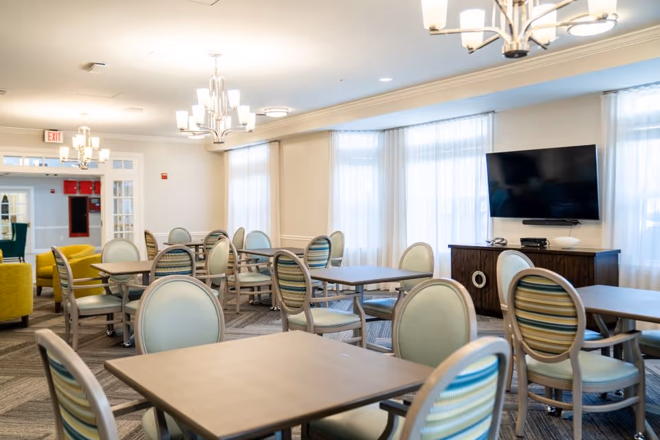 A bright and spacious dining area with multiple square tables and chairs featuring light blue and striped upholstery. The room has large windows with sheer white curtains, modern chandeliers, and a flat-screen TV mounted on the wall above a wooden cabinet. There are also yellow armchairs visible in the background near a doorway.