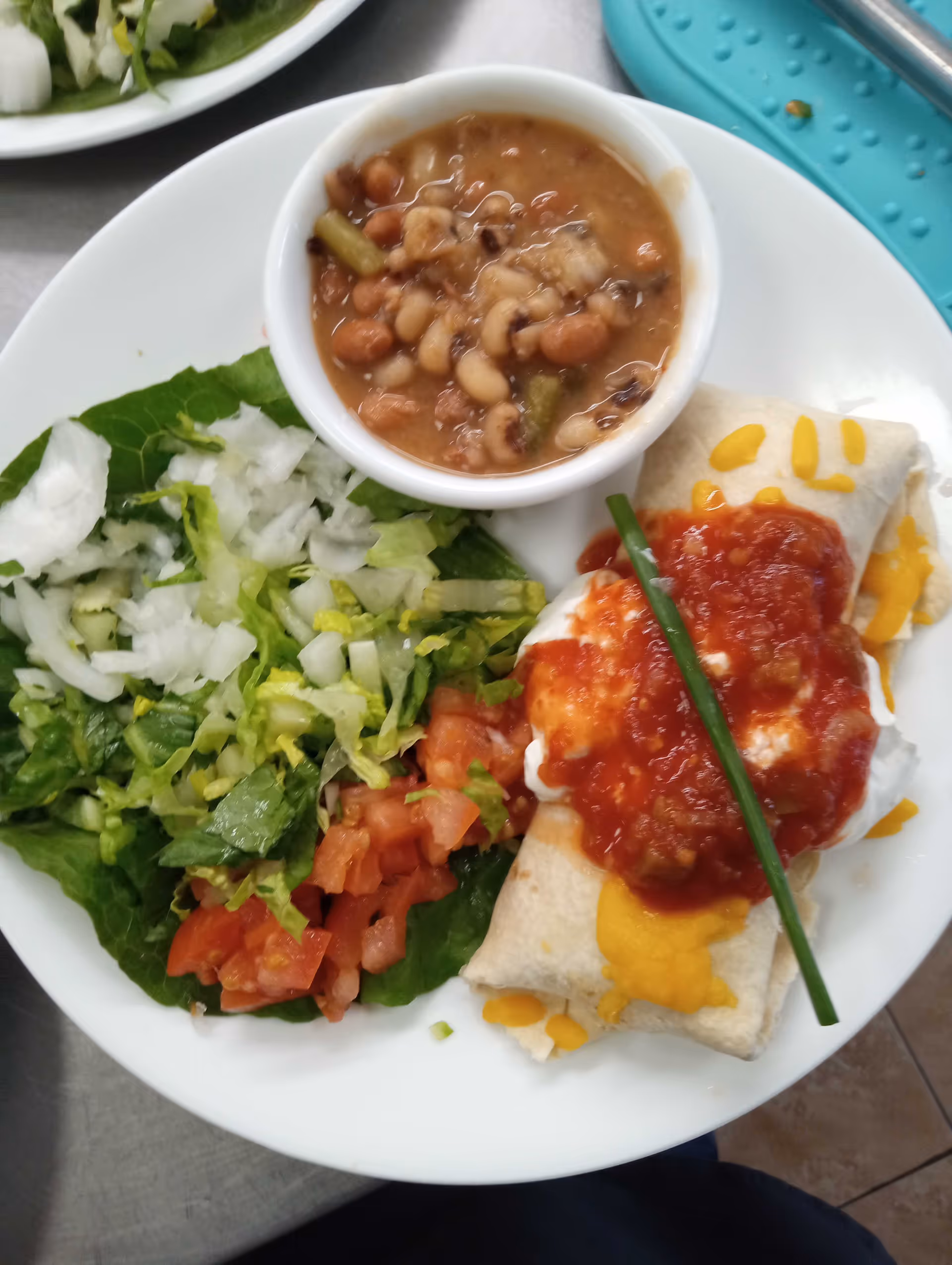 A white plate with a serving of mixed salad including lettuce, chopped onions, and diced tomatoes, a small bowl of mixed beans in broth, and a burrito topped with melted cheddar cheese, sour cream, salsa, and a green chive garnish.