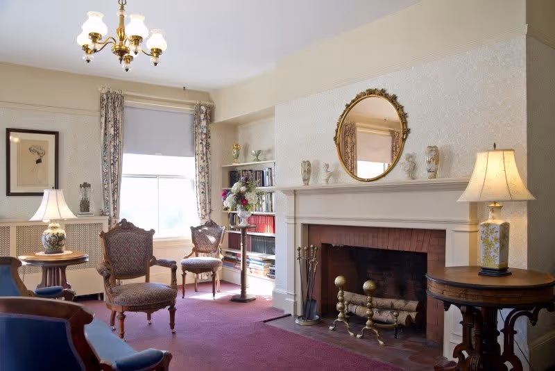 A cozy living room with antique-style furniture including upholstered chairs and wooden tables. There is a fireplace with brass andirons and logs, a round ornate mirror above the mantel, and decorative busts on the mantel. The room has floral curtains framing a window, a chandelier, and two table lamps providing warm lighting. Bookshelves with books and a vase of flowers are also visible.