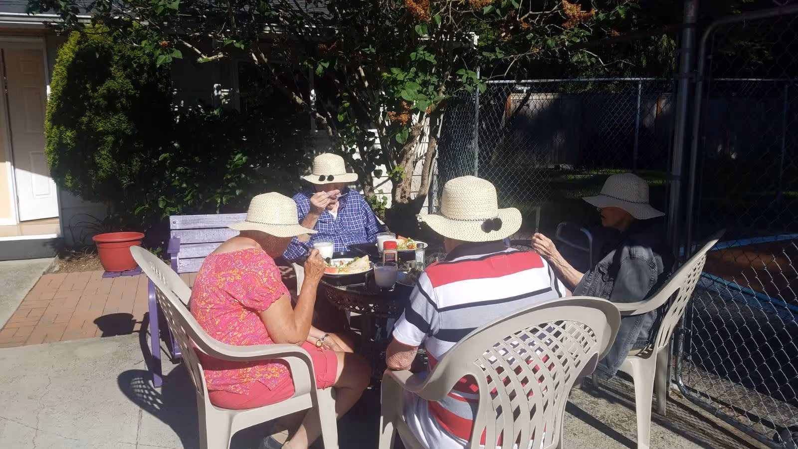Four elderly individuals wearing sun hats sitting around a small outdoor table enjoying a meal together on a sunny day. They are seated on plastic chairs and a purple bench, with a chain-link fence and greenery in the background.
