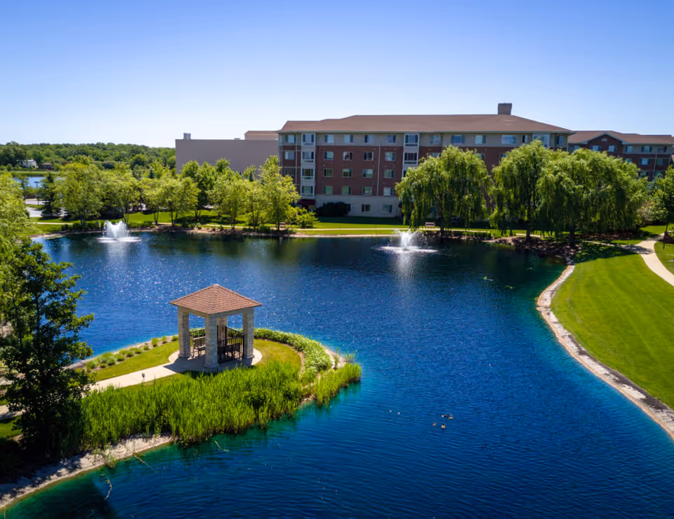 A scenic outdoor view of a senior living facility named Sedgebrook featuring a large pond with two water fountains, a small gazebo on a grassy island, lush green trees, and a multi-story building in the background under a clear blue sky.