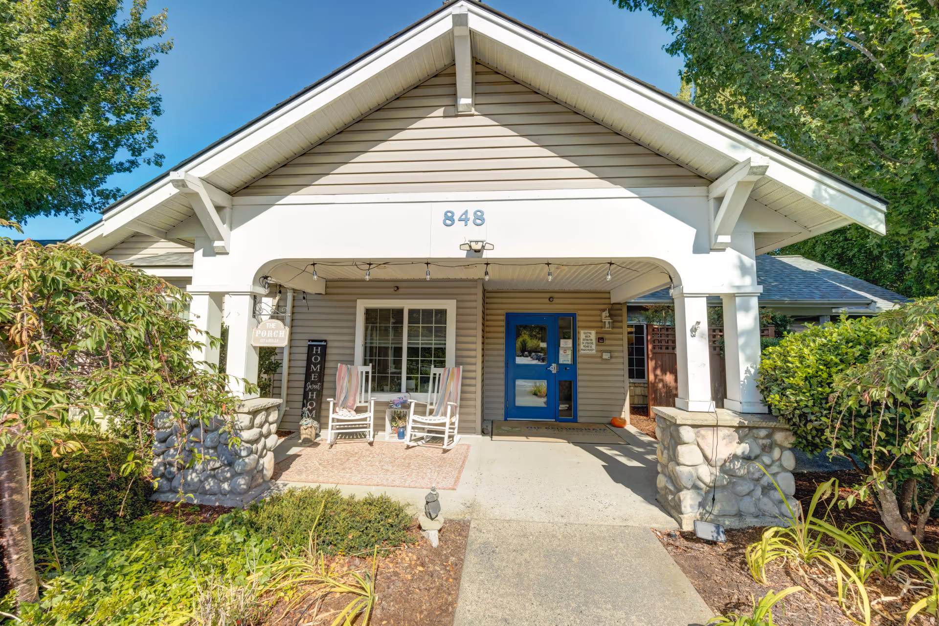 Front entrance of a senior living facility with a covered porch featuring two white rocking chairs with cushions and a small table with flowers. The building has beige siding, a blue door, and the number 848 above the entrance. There are plants and trees surrounding the walkway leading to the door.