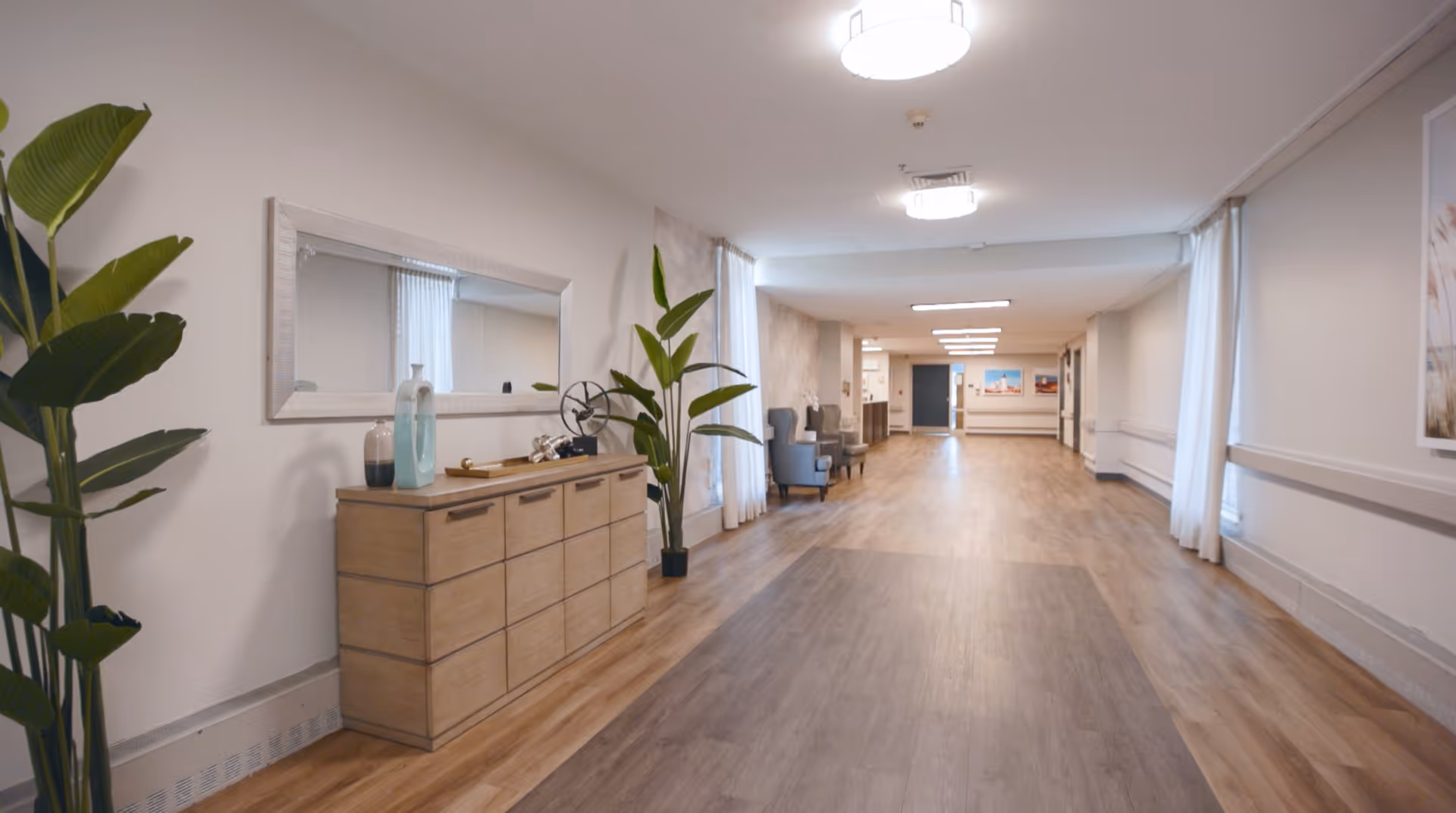 A wide, well-lit hallway in a senior living facility with wooden flooring, light-colored walls, and ceiling lights. On the left side, there is a wooden cabinet with decorative items and a large mirror above it, flanked by tall green plants. Further down the hallway, there are a few armchairs and framed pictures on the walls.