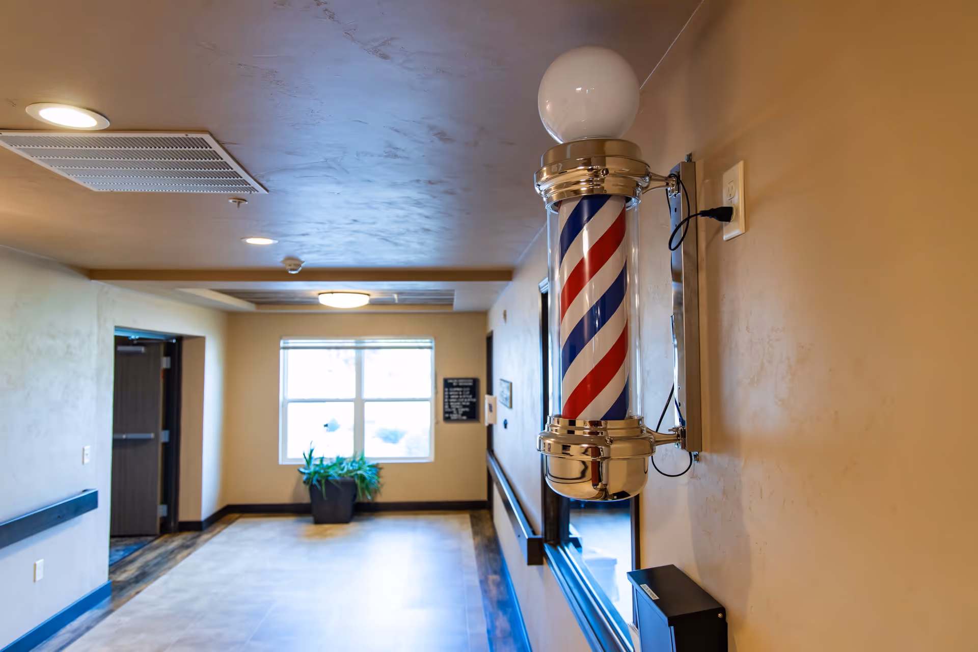 Interior hallway with a mounted barber pole on the right wall and a window with a potted plant at the far end.
