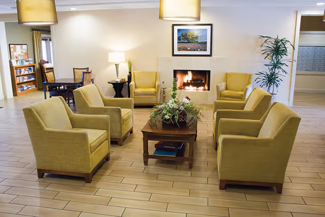 A cozy living room area with six yellow armchairs arranged around a wooden coffee table with a floral centerpiece. A fireplace with a fire burning is centered on the far wall, above which hangs a framed landscape painting. The room has light-colored tiled flooring, a tall potted plant to the right, and warm lighting from ceiling lamps and a table lamp.