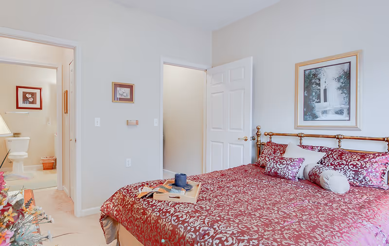 A bedroom with a large bed covered in a red and gold patterned bedspread and multiple pillows. There is a wooden tray with a cup and a book on the bed. The room has light-colored walls with two framed pictures hanging. An open door leads to a hallway, and another open door reveals a bathroom with a toilet and framed artwork on the wall.