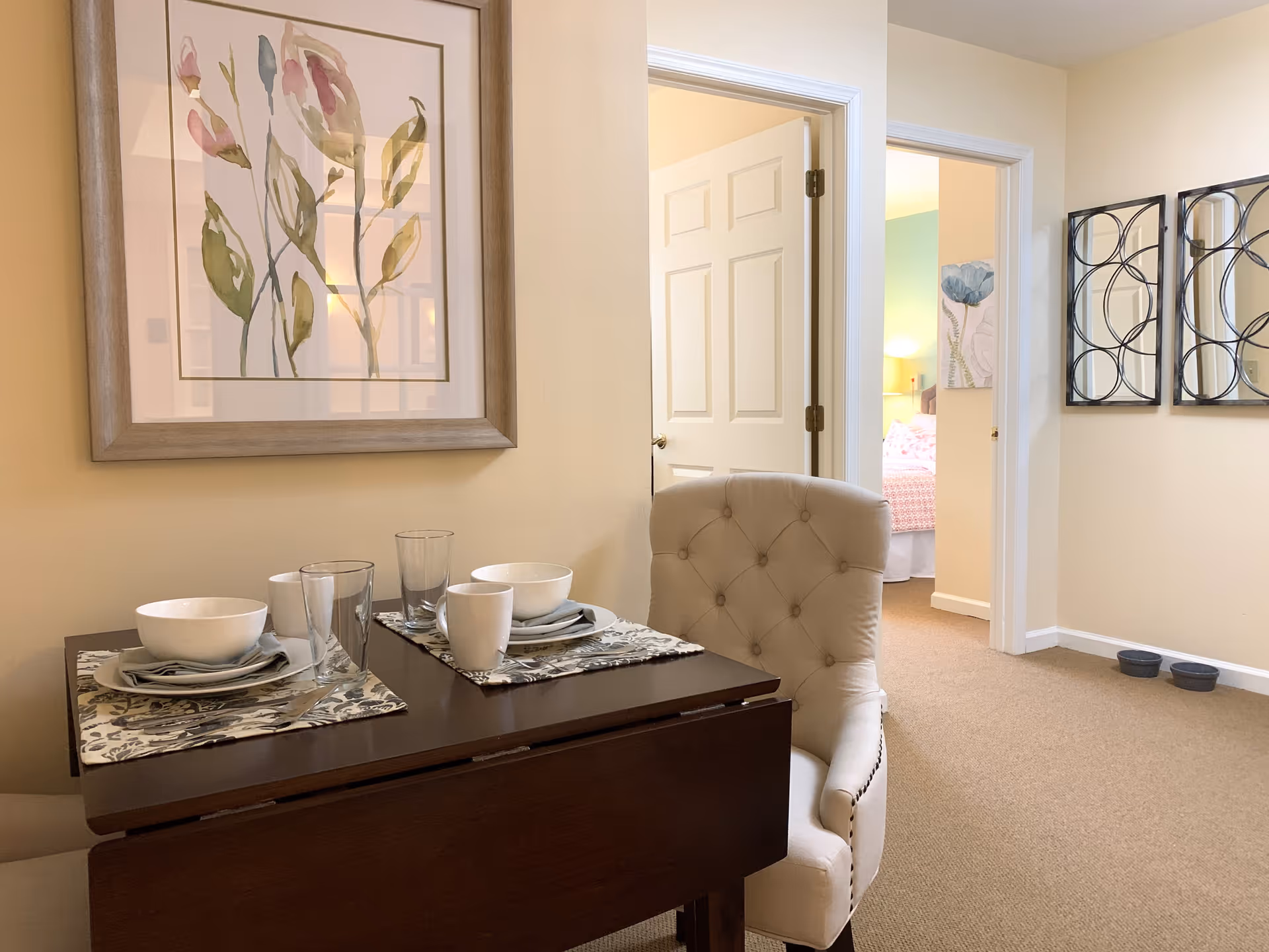 A small dining area with a dark wooden table set for two with white bowls, plates, cups, and glasses on patterned placemats. There are two beige upholstered chairs with button tufting. On the wall above the table is a framed floral painting. In the background, an open door reveals a bedroom with a bed and a bedside lamp. The walls are painted a soft beige color, and there are decorative mirrors on the adjacent wall.
