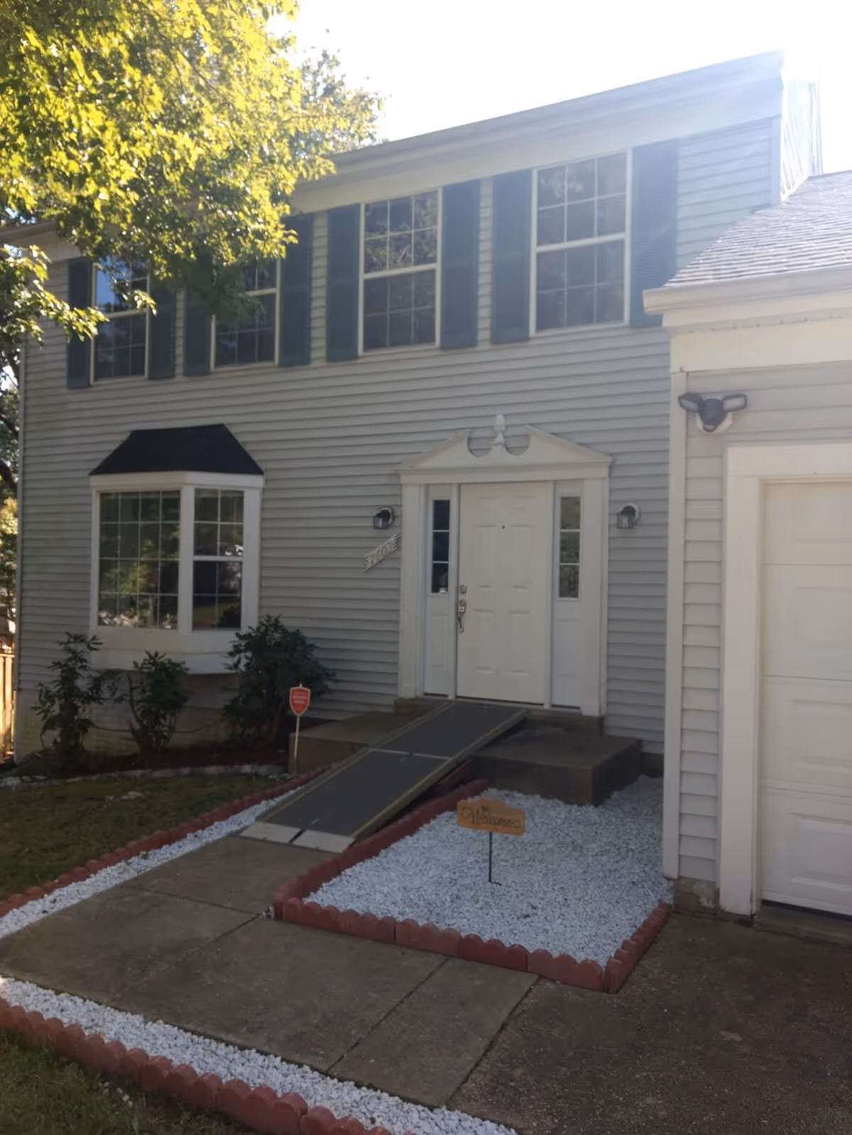 Front exterior view of a two-story house with light gray siding, white trim, and dark green shutters. There is a concrete walkway leading to the front door, which has a wheelchair ramp. The landscaping includes white gravel bordered by red bricks and some green bushes near a bay window. A small wooden sign with the word 'Welcome' is placed in the gravel area.