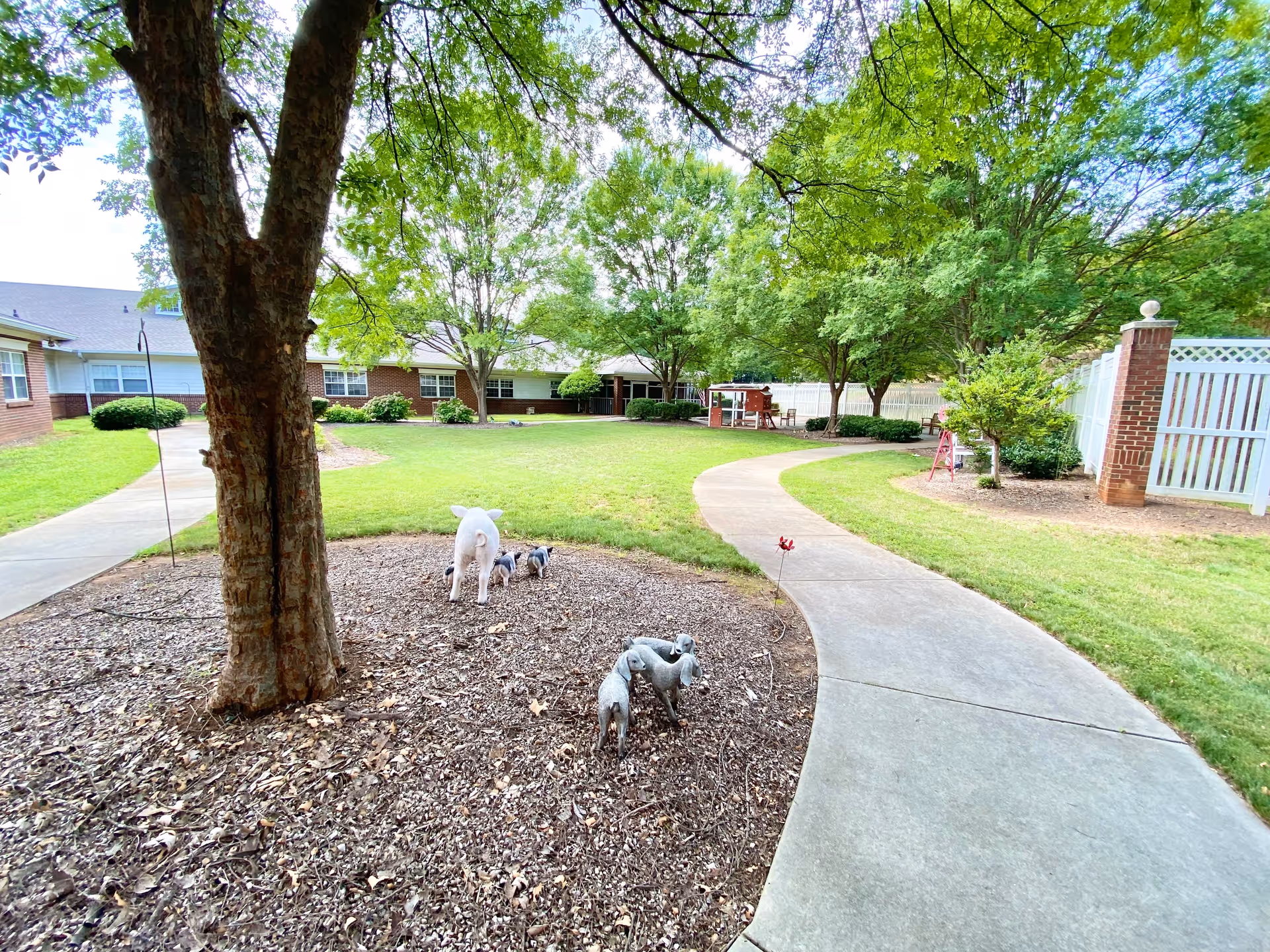 A peaceful outdoor courtyard area with a curved concrete walkway surrounded by green grass and trees. There are small decorative statues of dogs near the base of a tree, and a brick building with white fencing is visible in the background under a bright sky.
