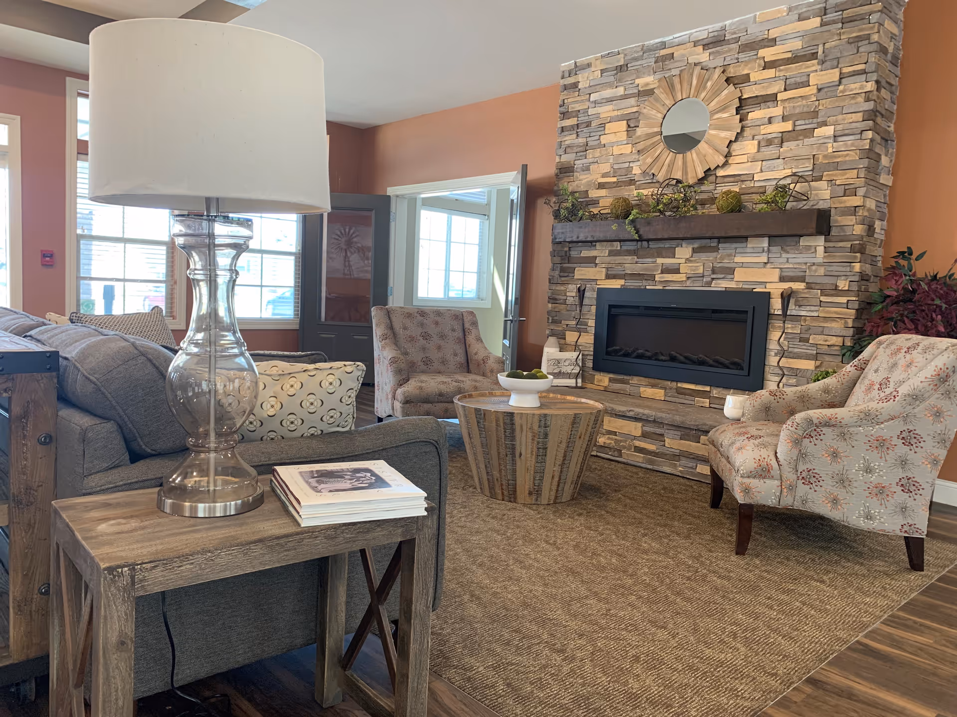 A cozy living room with a stone fireplace featuring a decorative round mirror and plants on the mantel. The room has two patterned armchairs, a wooden coffee table with a bowl of green apples, a gray sofa with patterned pillows, and a wooden side table with a clear glass lamp and stacked books. Large windows and a door with glass panels allow natural light to fill the space.