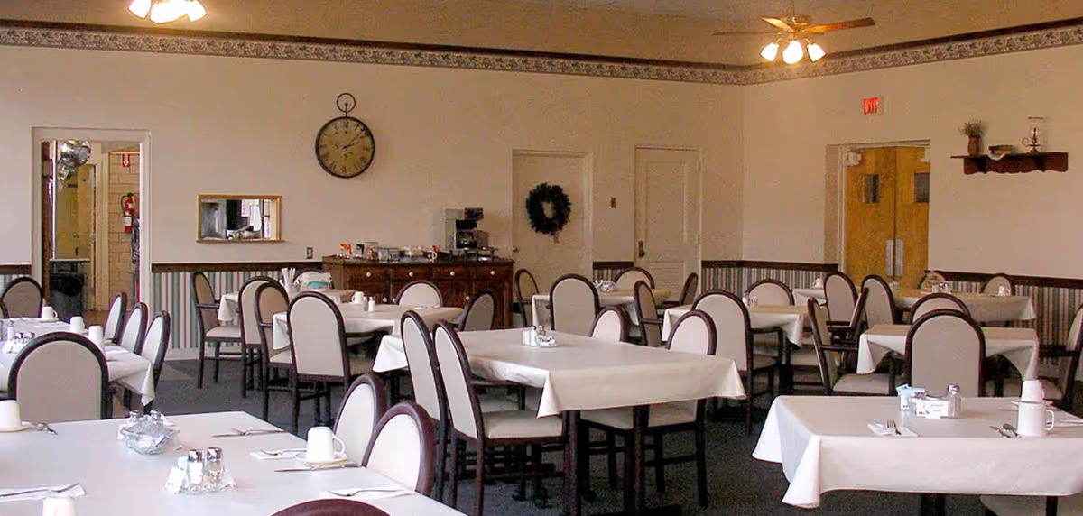 A dining room with multiple tables covered in white tablecloths and surrounded by chairs with light upholstery and dark frames. The room has a clock on the wall, a sideboard with a coffee machine and condiments, and ceiling fans with lights. There are doors and a window visible on the walls, along with some decorative elements like a wreath and a small shelf with items.