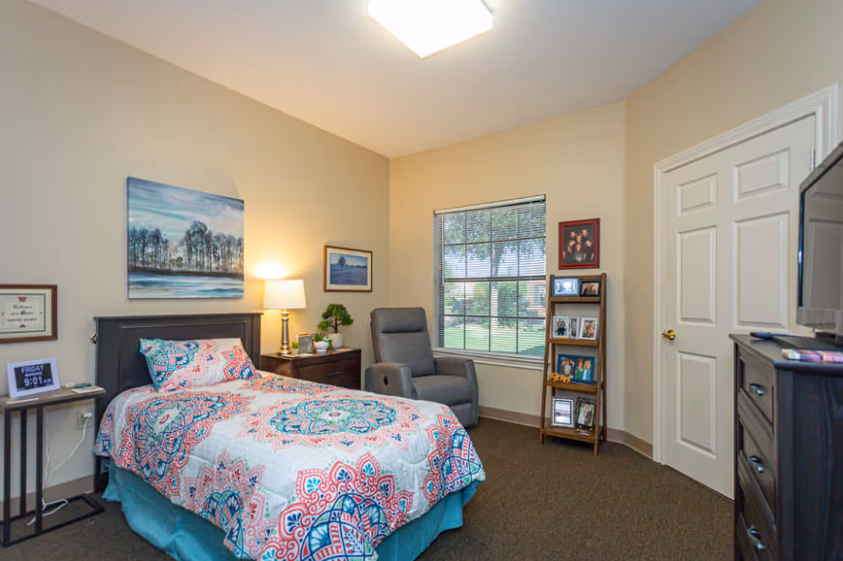 A cozy bedroom in a memory care facility featuring a single bed with colorful patterned bedding, a nightstand with a lamp and small plant, a comfortable gray recliner chair near a window with blinds, a small table with a digital clock, and a dresser with a TV on top. The walls are decorated with framed pictures and artwork.
