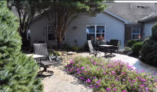 Outdoor patio area at Willow Brook Christian Village featuring a paved walkway, several chairs and tables, surrounded by green shrubs, trees, and purple flowering plants in front of a single-story building.