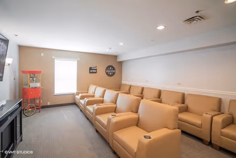 A small theater room with two rows of beige recliner chairs facing a large screen on the left wall. There is a red popcorn machine in the corner near a window with blinds. The walls are light-colored with a white chair rail molding, and the ceiling has recessed lighting.