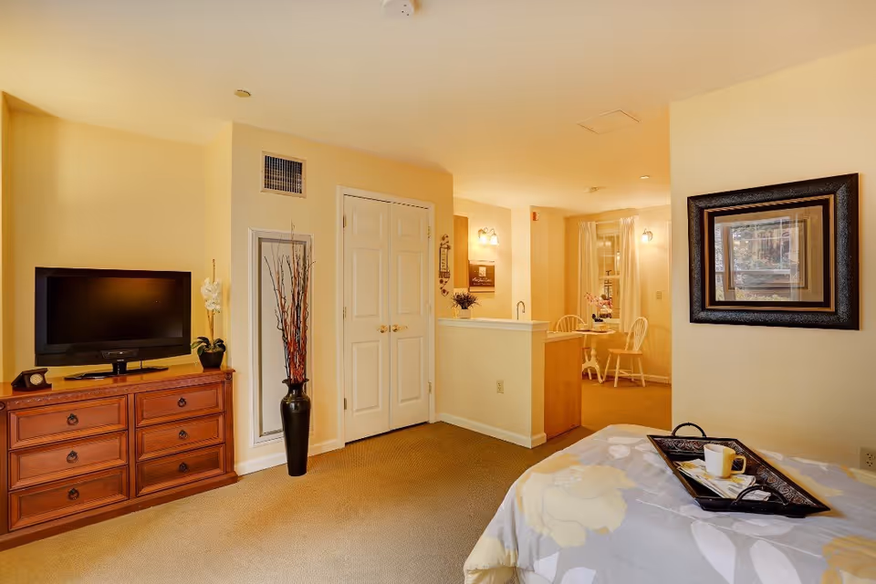 Studio-style interior with a bed and tray in the foreground, a TV on a dresser, and a small kitchenette/dining area in the background.