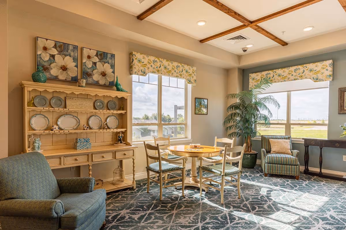 A cozy sitting area in an assisted living facility with a round wooden table surrounded by four chairs with light blue cushions. There is a blue upholstered armchair on the left and a striped armchair with a pillow near the window on the right. The room has large windows with floral valances letting in natural light, a wooden hutch displaying plates and decorative items, a potted plant, and framed artwork on the walls. The ceiling features wooden beams and recessed lighting.