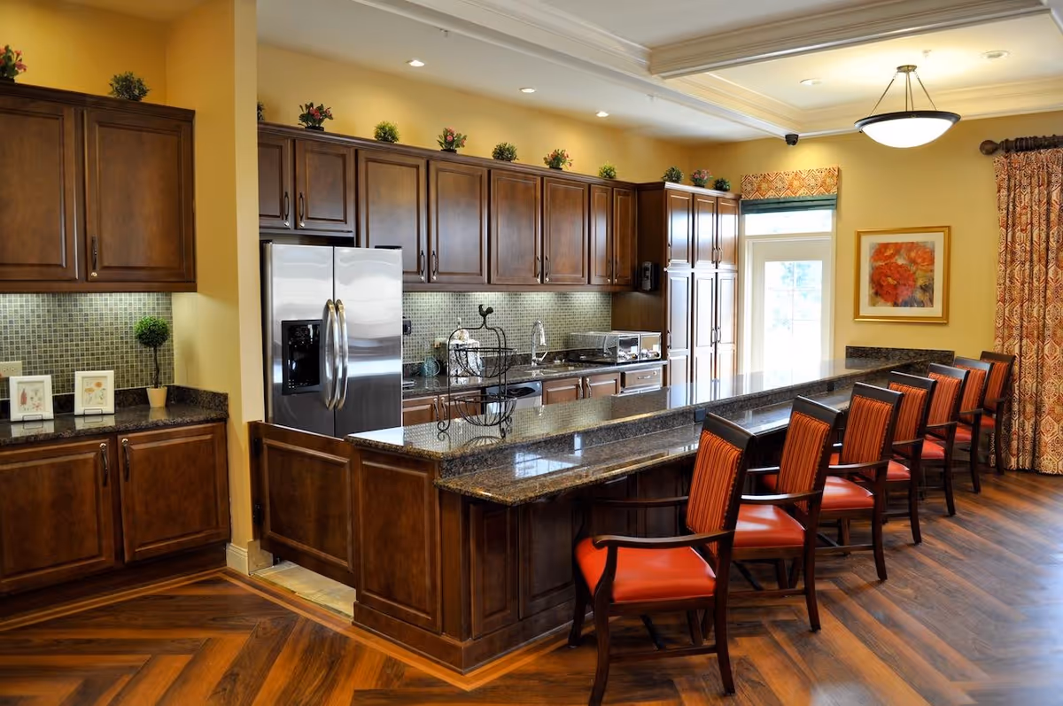 A spacious kitchen area with dark wood cabinets, a stainless steel refrigerator, and a long granite countertop with six red cushioned chairs. The room has warm yellow walls, a decorative light fixture on the ceiling, and a framed floral painting on the wall near a door with a window. The floor is wood with a herringbone pattern.