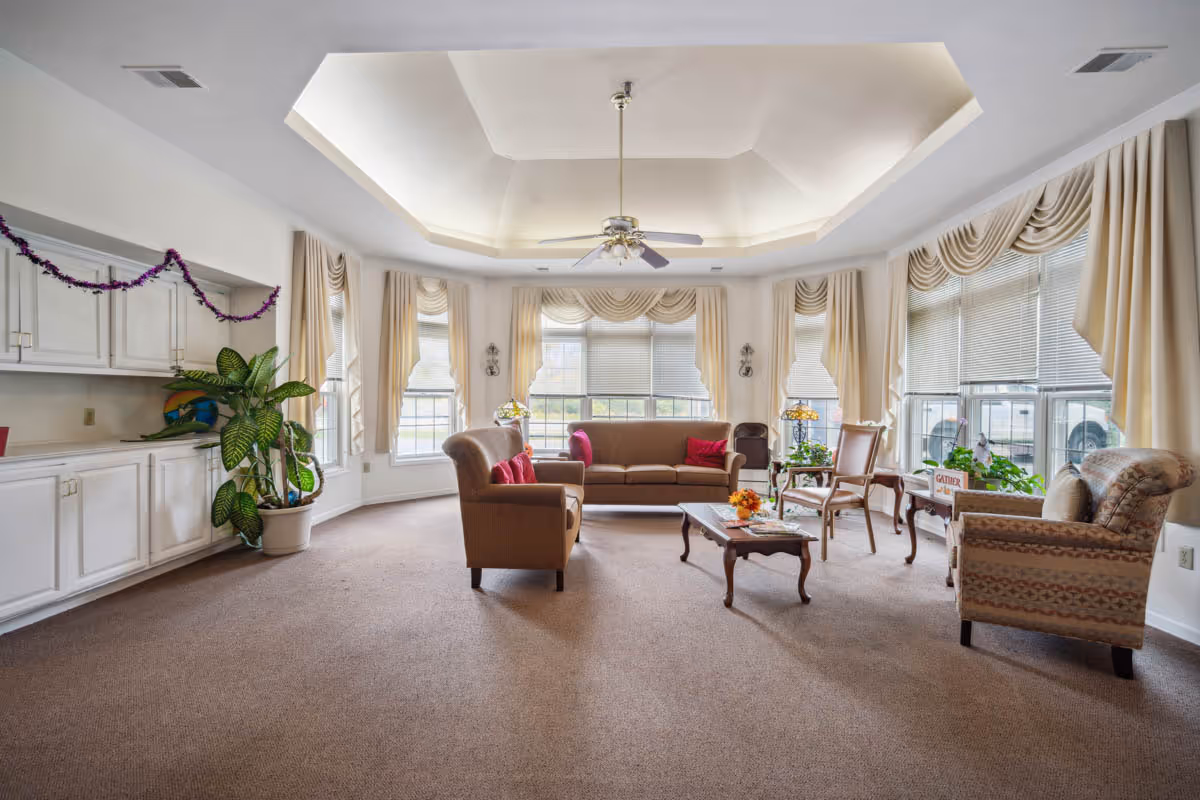 A spacious living room with beige carpet and a tray ceiling with a ceiling fan. The room has large windows with cream-colored curtains and blinds, allowing natural light to fill the space. There is a brown sofa with red cushions, two armchairs, a wooden coffee table with a small flower arrangement, and a side table with a lamp and plants. White cabinets line one wall, decorated with a purple garland and a large potted plant nearby.
