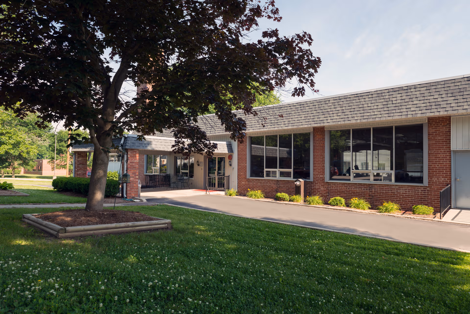 Exterior view of a single-story brick building with large windows and a shingled roof, surrounded by green grass and trees under a partly cloudy sky.