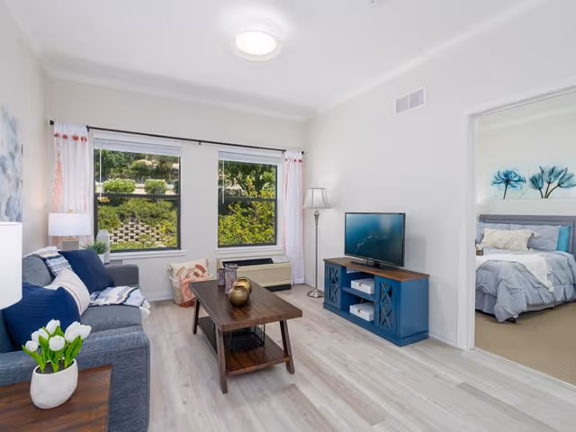 A bright living room with light wood flooring, a gray sofa with blue and white cushions, a wooden coffee table with decorative items, a blue TV stand with a flat-screen TV, a floor lamp, and two large windows with white and pink curtains showing greenery outside. An adjacent bedroom with a bed and blue floral wall art is visible through an open doorway.