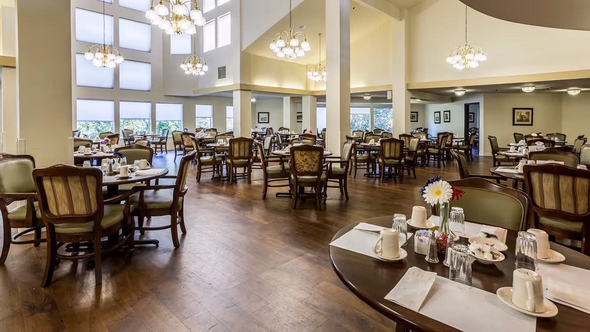 Spacious, well-lit dining room with multiple round tables set for meals, wooden chairs, chandeliers, and large windows.